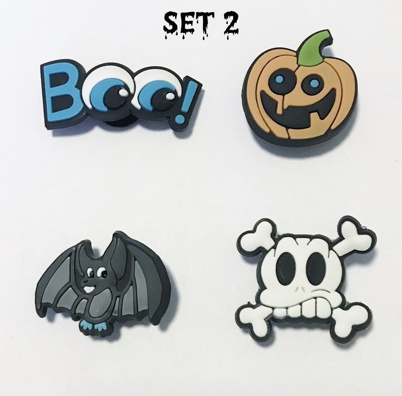 Op de afbeelding: Set van vier Halloween-schoenbedels. De bedels zijn een pompoen met een glimlachend gezicht, een vleermuis met blauwe voeten, een schedel met kruisbeenderen en een blauwe en witte "Boo!" bedel met oogjes.