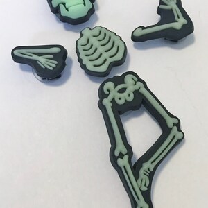 Op de afbeelding: Set met skeletcharms die in het donker gloeien, inclusief een schedel, ribbenkast en twee armen en benen. De charms zijn gemaakt van zwart plastic met een groene gloed-in-the-dark afwerking.