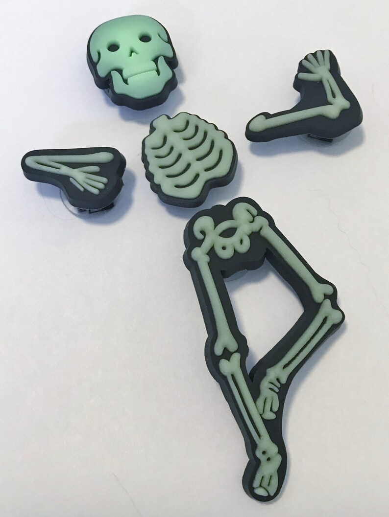 Op de afbeelding: Set met skeletcharms die in het donker gloeien, inclusief een schedel, ribbenkast en twee armen en benen. De charms zijn gemaakt van zwart plastic met een groene gloed-in-the-dark afwerking.