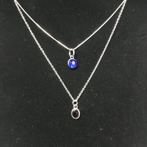 May include: A silver chain necklace with two pendants. The top pendant features a blue crystal, and the bottom pendant features a purple crystal.