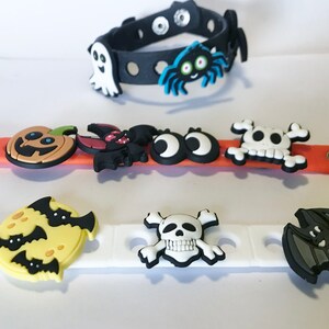 May include: Three silicone wristbands featuring Halloween-themed charms. The black band has a ghost and spider. The orange band features a pumpkin, bat, eyes, and skull. The white band displays a moon with bats, a skull, and a bat charm.