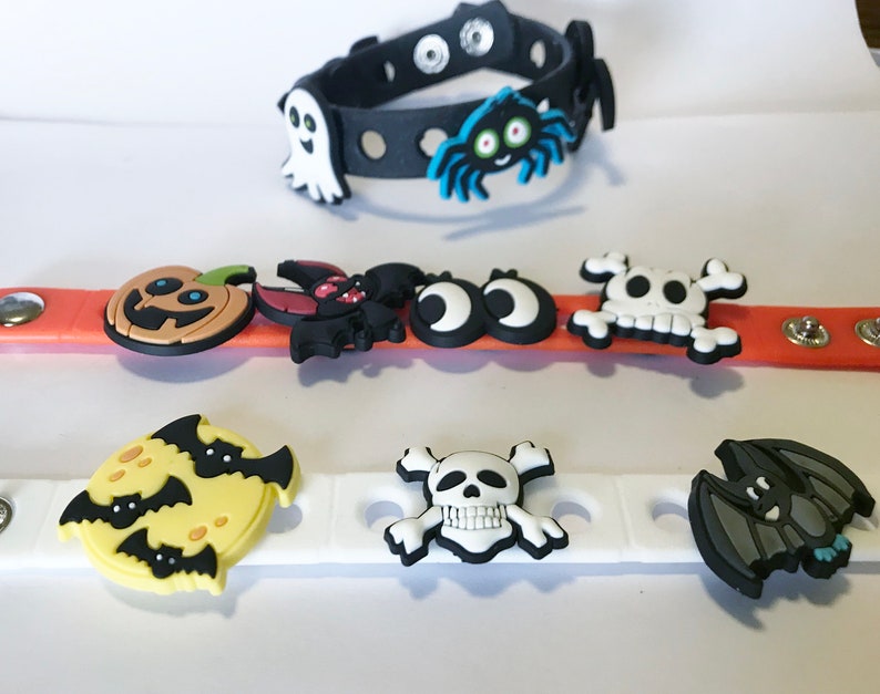 May include: Three silicone wristbands featuring Halloween-themed charms. The black band has a ghost and spider. The orange band features a pumpkin, bat, eyes, and skull. The white band displays a moon with bats, a skull, and a bat charm.