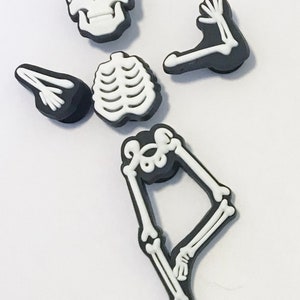 Op de afbeelding: Een set van vijf zwarte en witte skeletcharms. De charms zijn gevormd als een schedel, ribbenkast en twee armen en benen. De charms zijn gemaakt van plastic en hebben een glanzende afwerking.
