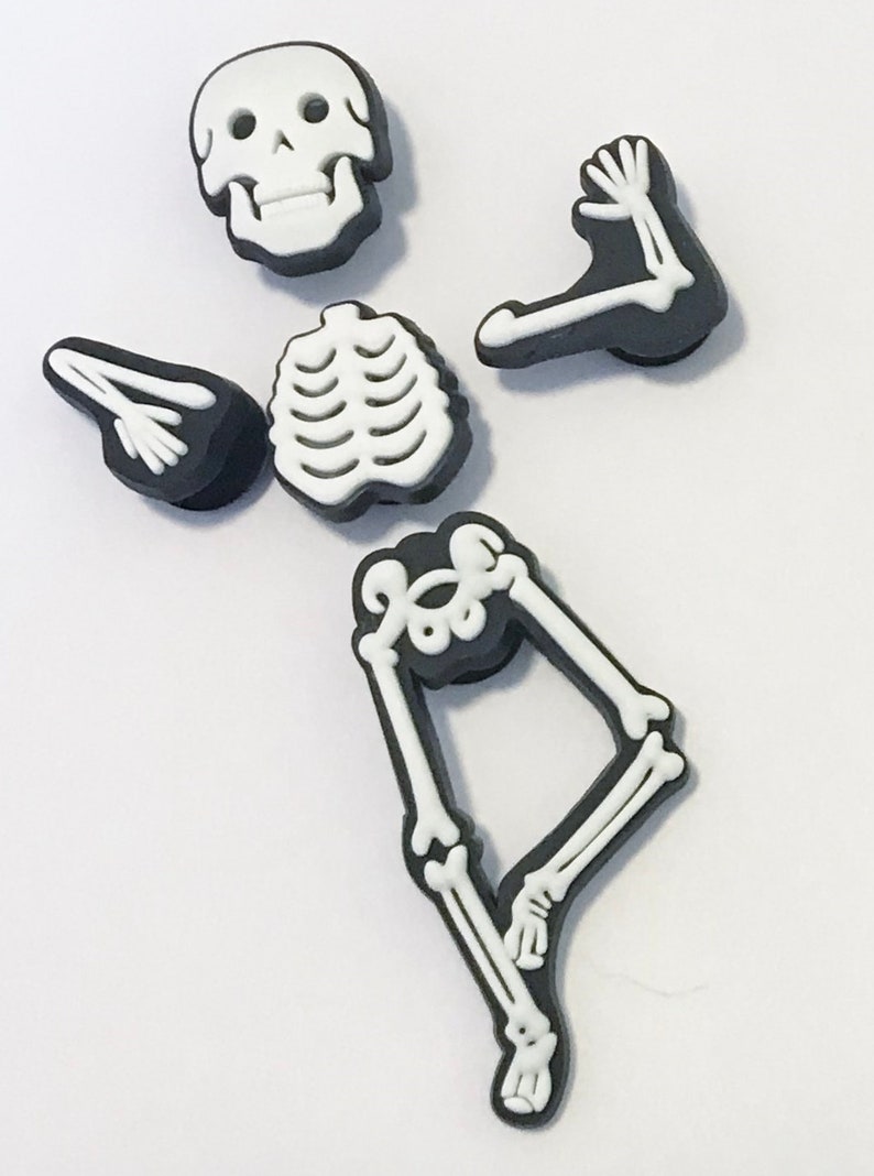 Op de afbeelding: Een set van vijf zwarte en witte skeletcharms. De charms zijn gevormd als een schedel, ribbenkast en twee armen en benen. De charms zijn gemaakt van plastic en hebben een glanzende afwerking.