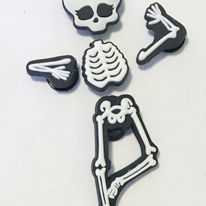 Op de afbeelding: Een set van vijf zwarte en witte skeletcharms met een roze strik op de schedel. De charms zijn voor een schoen of ander accessoire.