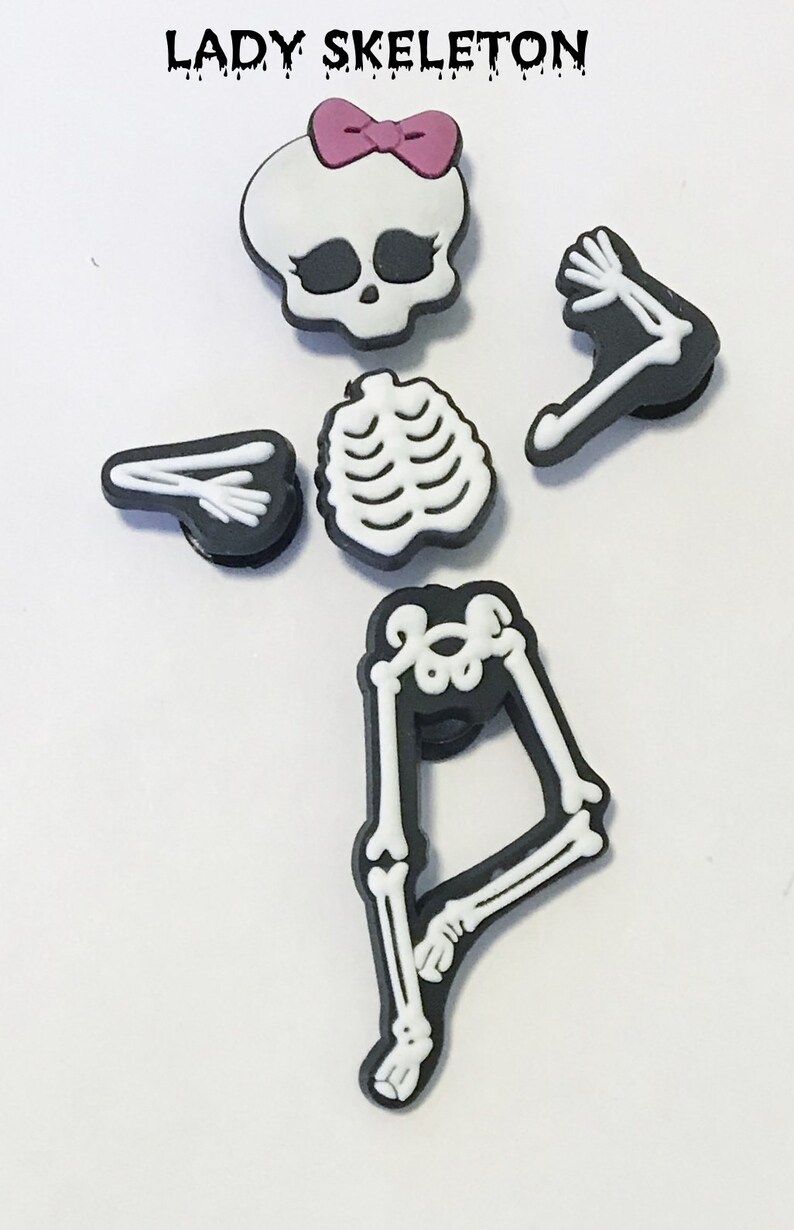 Op de afbeelding: Een set van vijf zwarte en witte skeletcharms met een roze strik op de schedel. De charms zijn voor een schoen of ander accessoire.