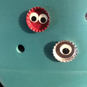 May include: Two bottle caps with googly eyes attached to a teal surface. One bottle cap is red and the other is silver.