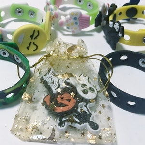 Puede incluir: Una colecci&oacute;n de pulseras de silicona a presi&oacute;n de colores con varios dijes decorativos, incluyendo una carita sonriente, una magdalena y figuras de Halloween. Una peque&ntilde;a bolsa con ribete dorado contiene dijes adicionales.