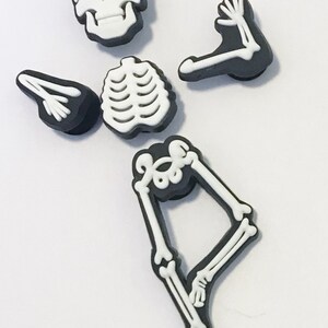 May include: A set of five black and white shoe charms shaped like individual skeleton parts, including a skull, a rib cage, two arm bones, and a leg bone piece. Each piece features a raised white bone design on a solid black background.