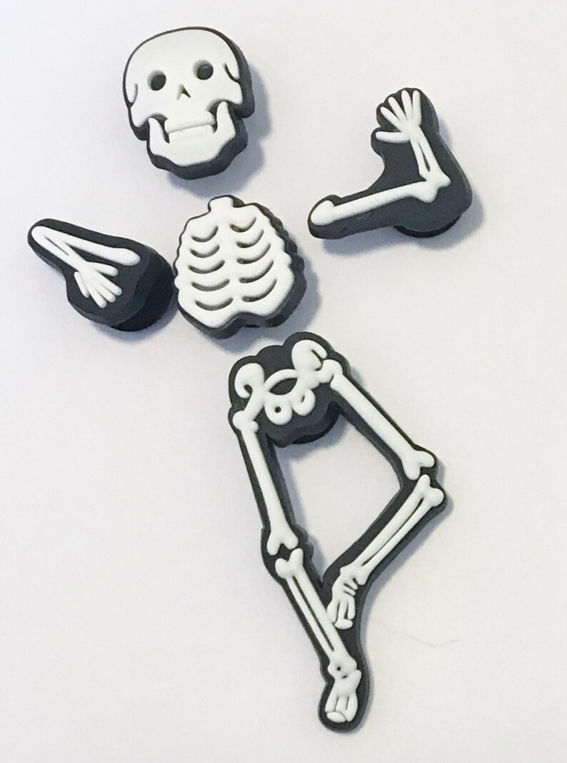 May include: A set of five black and white shoe charms shaped like individual skeleton parts, including a skull, a rib cage, two arm bones, and a leg bone piece. Each piece features a raised white bone design on a solid black background.