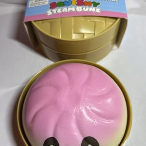 May include: A pink and yellow squishy toy shaped like a steamed bun, with black eyes and a smiling mouth. The toy is in a gold-colored container, with a cardboard lid that reads "Dim Sum" and "Squishy Steam Bunz".