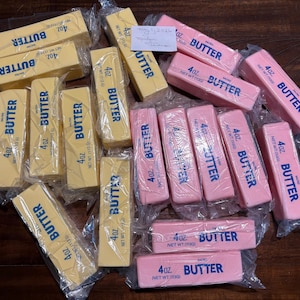 Könnte beinhalten: Eine Ansammlung von verpackten Butterstangen, gelb und rosa, liegt auf einer dunklen Holzoberfläche. Jede Stange ist mit dem Wort "BUTTER" und dem Gewicht, 113g, beschriftet. Die Verpackung ist aus klarem Kunststoff.