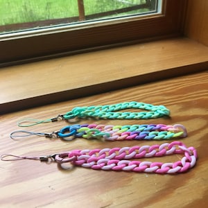 May include: Three colorful phone charms with a chain link design. The charms are in shades of green, pink, and rainbow.