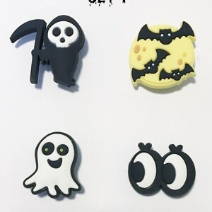 Op de afbeelding: Set van vier Halloween-schoenbedels. De bedels zijn zwart, wit en geel en bevatten een Grim Reaper, een geest, een maan met vleermuizen en twee oogjes.