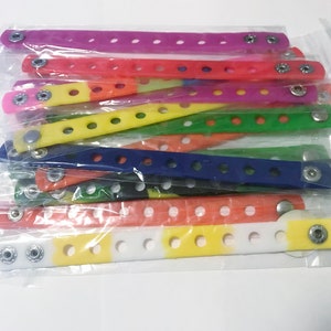 Puede incluir: Una colecci&oacute;n de pulseras de mu&ntilde;eca coloridas con cierre de bot&oacute;n a presi&oacute;n, en varios colores: morado, rojo, amarillo, verde, azul y naranja. Cada pulsera est&aacute; hecha de un material flexible y tiene m&uacute;ltiples agujeros para ajustar. Las pulseras est&aacute;n empaquetadas en pl&aacute;stico transparente.