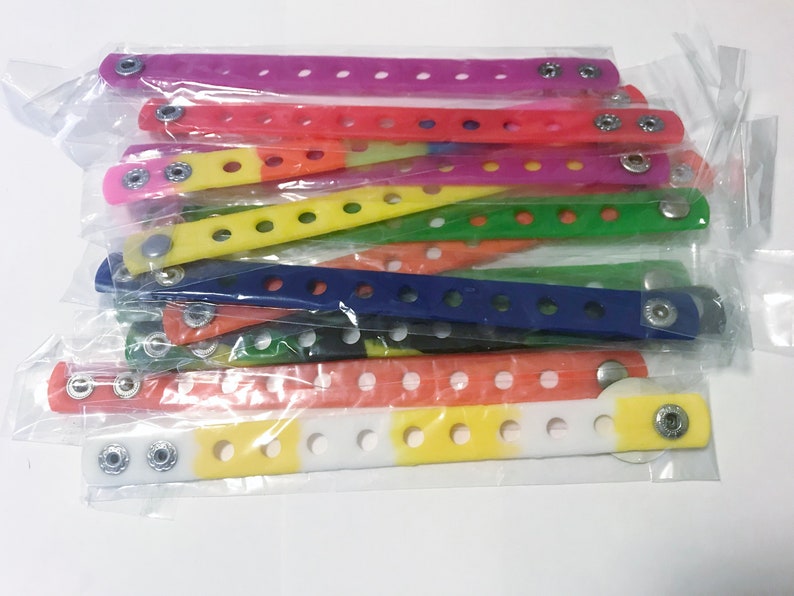 Puede incluir: Una colecci&oacute;n de pulseras de mu&ntilde;eca coloridas con cierre de bot&oacute;n a presi&oacute;n, en varios colores: morado, rojo, amarillo, verde, azul y naranja. Cada pulsera est&aacute; hecha de un material flexible y tiene m&uacute;ltiples agujeros para ajustar. Las pulseras est&aacute;n empaquetadas en pl&aacute;stico transparente.