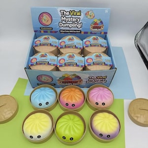 Puede incluir: Una caja expositora de juguetes "The Viral Mystery Dumpling!". La caja contiene varios juguetes con forma de dumpling en varios colores, incluyendo azul, naranja, rosa, amarillo y verde. Cada dumpling tiene una cara sonriente y viene en un pequeño recipiente redondo.