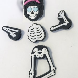 Op de afbeelding: Een set van vijf zwarte en witte skeletcharms met een roze hoofdband. De charms omvatten een schedel met een zonnebril, een ribbenkast, twee armen en een paar benen.