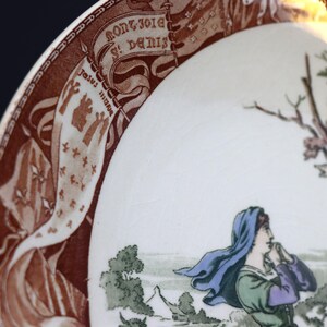 Puede incluir: Plato de cer&aacute;mica vintage con una ilustraci&oacute;n detallada. El plato presenta un borde marr&oacute;n con texto y elementos decorativos. La imagen central representa una figura con un velo azul, con un fondo de paisaje. El plato tiene un interior blanco.