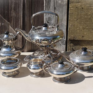 Op de afbeelding: Een complete zilveren theeset, inclusief een theepot, melkkan, suikerpot en serveerschalen. De set heeft een reflecterende, gepolijste afwerking en sierlijke details. De theepot wordt ondersteund door een decoratieve standaard.