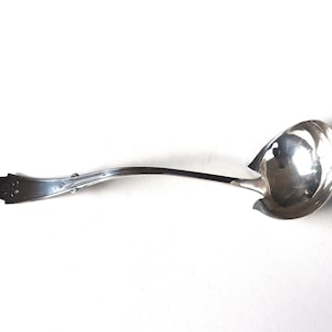 Puede incluir: Un cucharón de plata con un mango largo y curvo. El mango tiene un diseño decorativo en el extremo.