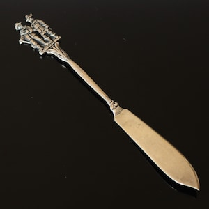 Puede incluir: Cuchillo de mantequilla plateado con un mango decorativo que representa tres figuras con vestimenta tradicional. La hoja es ancha y curvada, con un acabado pulido. Un utensilio de servicio clásico.