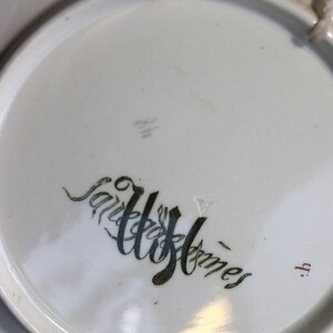 Puede incluir: Un plato de cer&aacute;mica blanco con las palabras "Sairee Homes" en una escritura estilizada. El plato tiene un dise&ntilde;o sencillo y un agujero para colgarlo. El plato es redondo y tiene un borde ligeramente elevado.