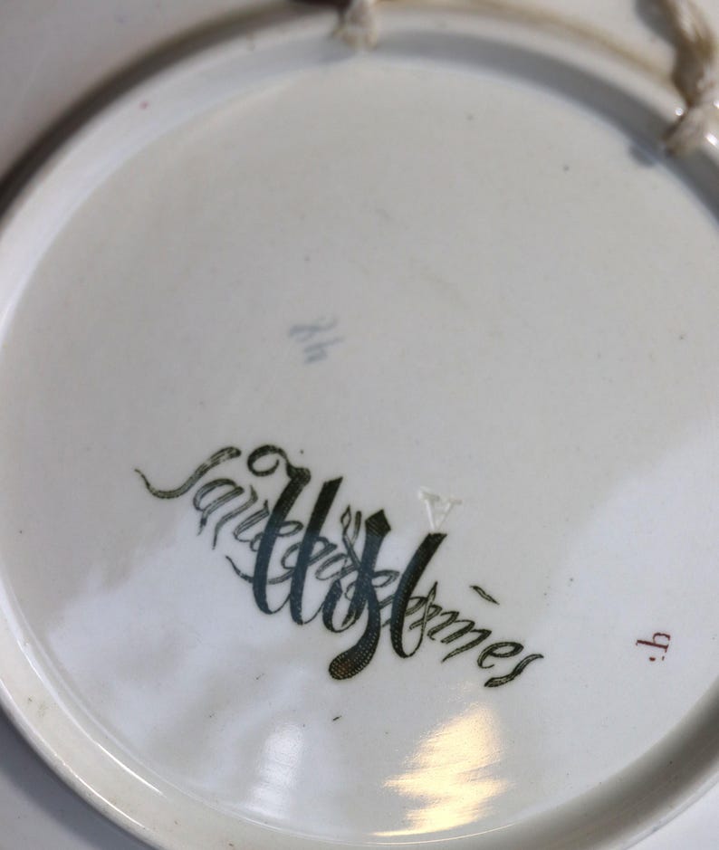 Puede incluir: Un plato de cer&aacute;mica blanco con las palabras "Sairee Homes" en una escritura estilizada. El plato tiene un dise&ntilde;o sencillo y un agujero para colgarlo. El plato es redondo y tiene un borde ligeramente elevado.