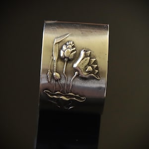 May include: A silver-toned cuff bracelet featuring a detailed embossed design of lotus flowers and leaves. The floral elements are raised against a textured background, creating a three-dimensional effect. The bracelet has a rectangular shape.