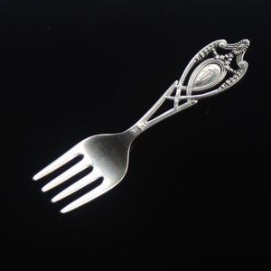May include: A silver baby fork with an ornate handle. The handle has a decorative design with a small shield in the center.