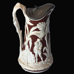 May include: A dark reddish-brown ceramic pitcher featuring white relief carvings of palm leaves and a figure carrying another person on their back. The pitcher has a textured surface, a curved handle, and a dark metallic hinged lid at the top.