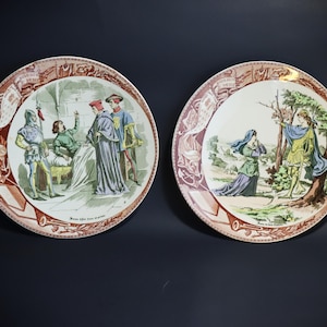 Puede incluir: Dos platos decorativos con ilustraciones de la &eacute;poca renacentista. Uno representa figuras en una habitaci&oacute;n, el otro un hombre y una mujer al aire libre. Los platos tienen un borde rojo y marr&oacute;n.