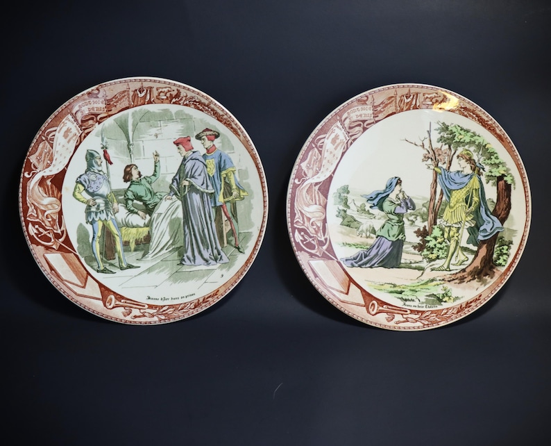 Puede incluir: Dos platos decorativos con ilustraciones de la &eacute;poca renacentista. Uno representa figuras en una habitaci&oacute;n, el otro un hombre y una mujer al aire libre. Los platos tienen un borde rojo y marr&oacute;n.