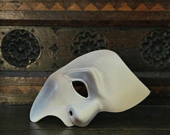 May include: A white, asymmetrical half-face mask with a smooth, matte finish. The mask features a single eye opening on the left side and a contoured nose section, covering only the right side of the face with a curved, sweeping edge design.