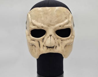 May include: A beige, bone-colored skull mask featuring deep, dark eye sockets and a prominent nasal cavity. The mask has a weathered, textured finish with subtle brown shading in the crevices, displayed on a black mannequin head form.