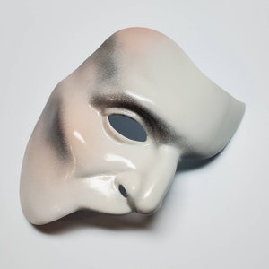 Puede incluir: Una m&aacute;scara blanca del Fantasma de la &Oacute;pera con un acabado liso y brillante. La m&aacute;scara tiene una nariz prominente y una sola abertura para los ojos. Presenta sutiles sombras en tonos grises y rosas, creando un efecto dram&aacute;tico. La m&aacute;scara est&aacute; dise&ntilde;ada para cubrir la mitad de la cara.