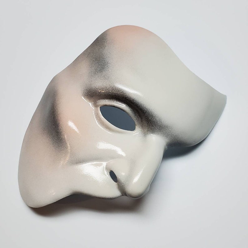 Puede incluir: Una m&aacute;scara blanca del Fantasma de la &Oacute;pera con un acabado liso y brillante. La m&aacute;scara tiene una nariz prominente y una sola abertura para los ojos. Presenta sutiles sombras en tonos grises y rosas, creando un efecto dram&aacute;tico. La m&aacute;scara est&aacute; dise&ntilde;ada para cubrir la mitad de la cara.