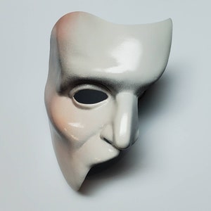 Puede incluir: Una m&aacute;scara blanca del Fantasma de la &Oacute;pera con un acabado liso y brillante. La m&aacute;scara tiene un dise&ntilde;o curvo, que cubre la mitad de la cara, con una nariz prominente y una sola abertura para el ojo. Los bordes est&aacute;n sombreados con negro y rosa.