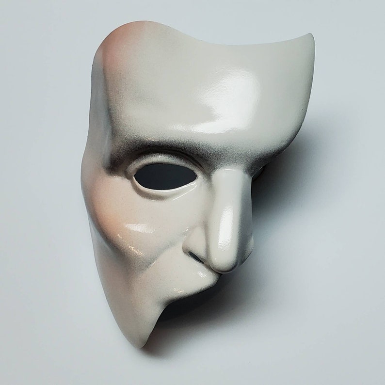Puede incluir: Una m&aacute;scara blanca del Fantasma de la &Oacute;pera con un acabado liso y brillante. La m&aacute;scara tiene un dise&ntilde;o curvo, que cubre la mitad de la cara, con una nariz prominente y una sola abertura para el ojo. Los bordes est&aacute;n sombreados con negro y rosa.