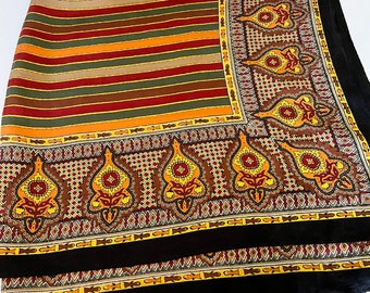 May include: A folded silk scarf with a striped pattern in shades of orange, brown, green, and gold. The scarf features a decorative border with ornate floral and geometric designs in brown, yellow, and red, framed by a black border.