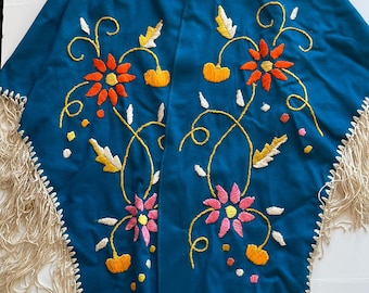 May include: A teal-colored shawl with embroidered floral designs in orange, pink, and yellow. The shawl has a collar and is trimmed with a cream-colored fringe along the bottom edge. The embroidery features detailed floral patterns.