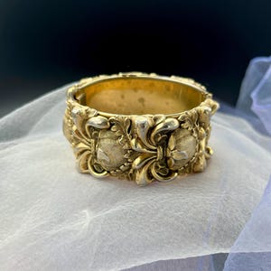 May include: A gold-colored, ornate bangle bracelet. The bracelet features detailed floral and shield-like designs. The metal has a polished finish, and the bracelet appears to be vintage or antique. The bracelet is displayed on a white, sheer fabric.