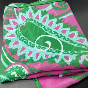 May include: A lightweight square scarf featuring a vibrant paisley pattern in shades of magenta, bright green, and light turquoise. The fabric has a semi-sheer quality with a finished green hem along the edges, displaying a repeating floral motif.