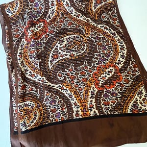 May include: A rectangular scarf with a brown border and a detailed paisley pattern in shades of brown, orange, white, and purple. The scarf has a silky texture and a dark brown border.