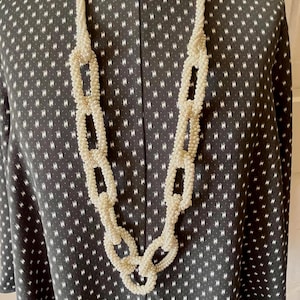 May include: A long, off-white beaded necklace with a chain-link design. The necklace is draped over a dark gray dress with a white dotted pattern. The necklace is made of small, round beads.