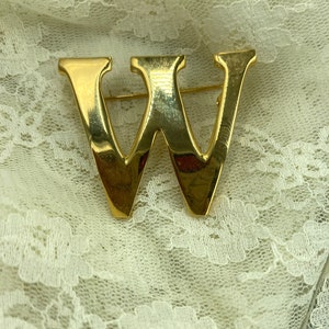 May include: A gold-colored metal brooch in the shape of the letter "W". The brooch has a pin on the back for attachment. The brooch is set against a white lace background.