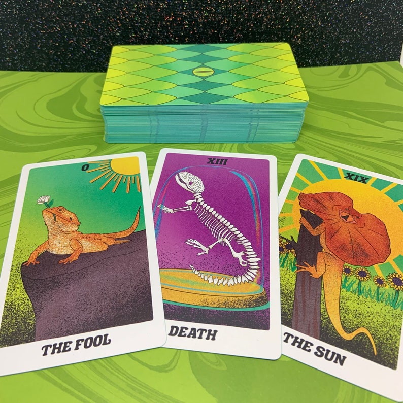 May include: Three tarot cards with colorful illustrations. The Fool card features a lizard with a flower on its head, the Death card shows a skeleton lizard, and the Sun card depicts a lizard with a frill.