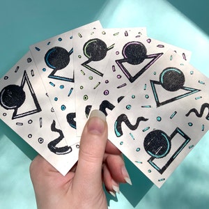 May include: Five white cards with black and colored geometric shapes and patterns. Each card features a black circle with a white outline and a different colored geometric shape around it. The cards are held in a hand.