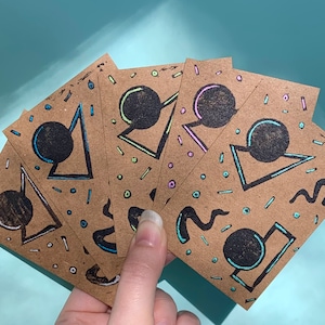 May include: Five brown cards with geometric shapes and black circles drawn on them. The shapes are outlined in neon green, pink, and blue. The cards are arranged in a fan shape.
