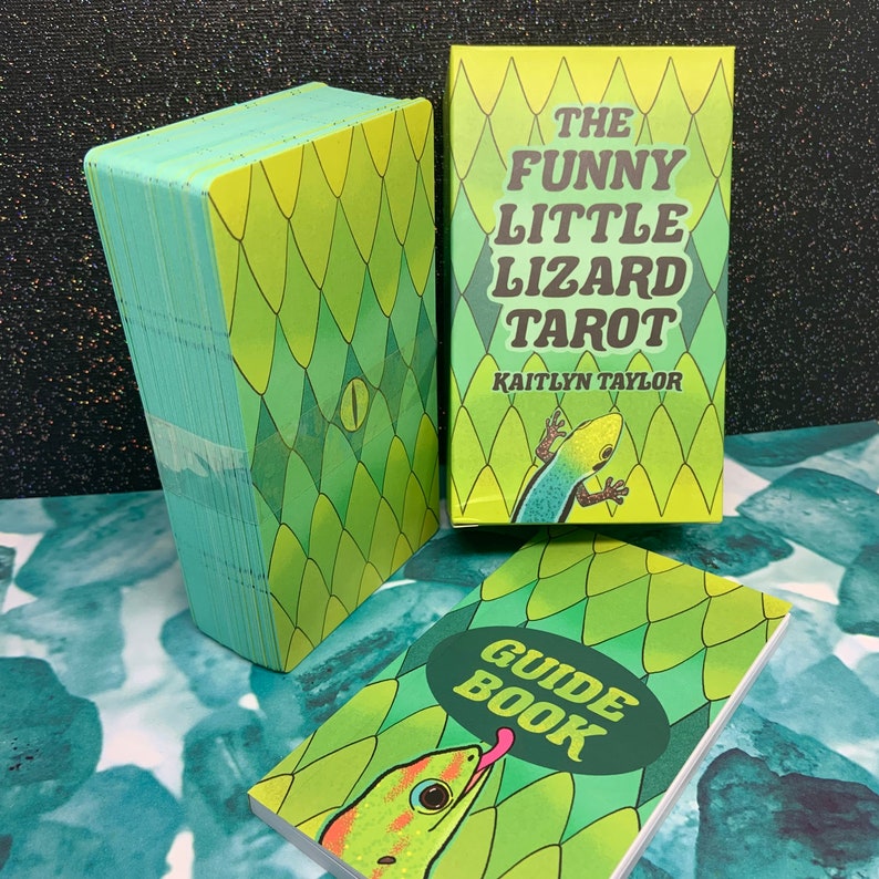 May include: A green and yellow tarot deck box with the title "The Funny Little Lizard Tarot" and the name "Kaitlyn Taylor". The box is next to a green and yellow guide book with the title "Guide Book".